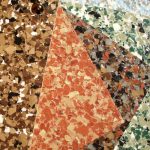 Close-up of a terrazzo flooring surface showing a section with a diagonal division where different colored stone chips are embedded in the concrete, featuring shades of brown, orange, black, white, and green