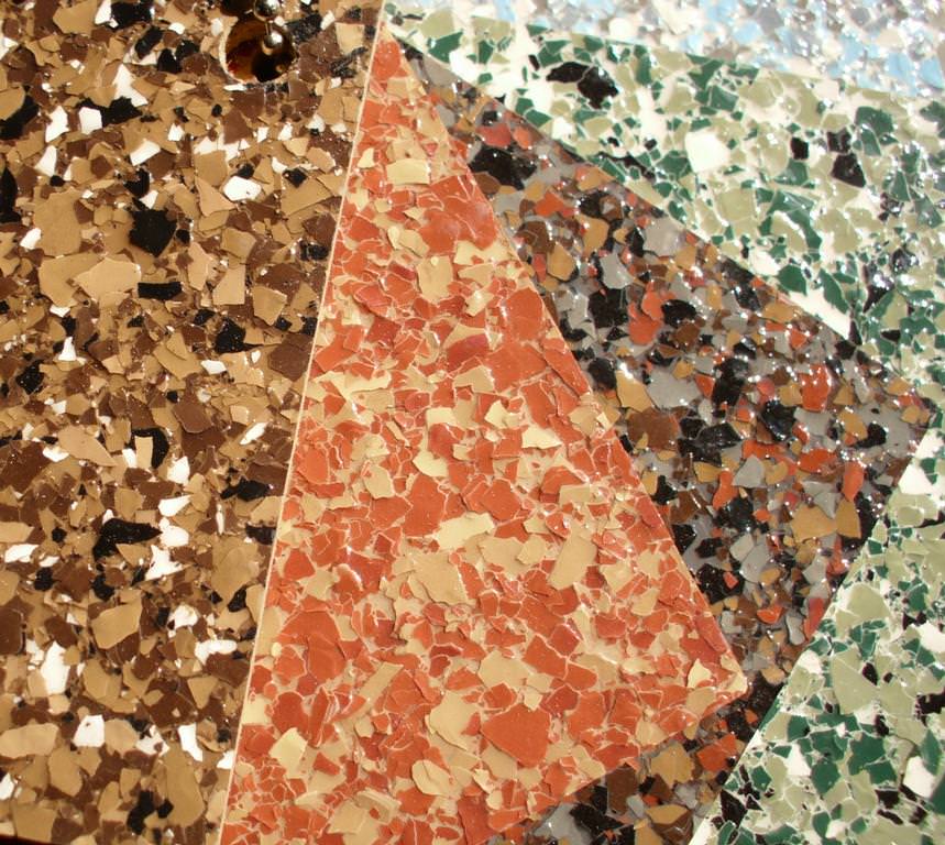 Close-up of a terrazzo flooring surface showing a section with a diagonal division where different colored stone chips are embedded in the concrete, featuring shades of brown, orange, black, white, and green