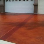 A wide driveway with a reddish-brown stained concrete finish, leading to a white garage door of a residential home. The smooth, uniformly colored surface is sectioned by control joints and features a manhole cover, highlighting a clean and well-maintained approach.