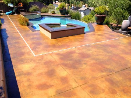 Luxurious backyard with a stained concrete pool deck in warm earth tones, leading to an inviting blue swimming pool with an integrated hot tub. The space is complemented by lush greenery, potted plants, and outdoor furniture, offering a relaxing outdoor retreat.