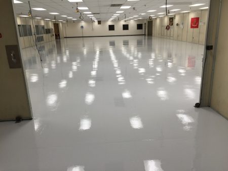 Spacious interior of an industrial building with a reflective white epoxy floor, featuring several hanging cables and emblems on distant walls under fluorescent lighting.