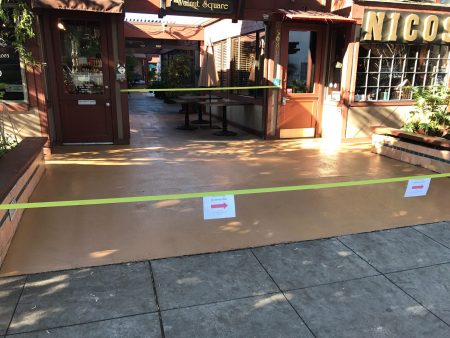 Outdoor view of a walkway with a freshly coated epoxy floor, cordoned off with yellow caution tape and signs, leading to a restaurant entrance with brown doors and green foliage.