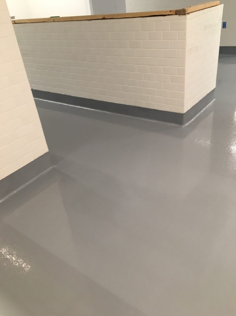 A corner of a room with walls composed of clean white tiles topped with a thin black stripe, meeting a glossy gray epoxy floor that reflects the lights above.