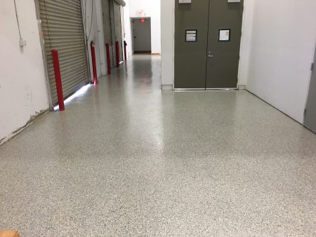 A clean and shiny terrazzo floor in a corridor with white walls. To the left, there are closed rolling shutter doors with red protective bollards at their base, and straight ahead, a pair of closed double doors with windows and signs.
