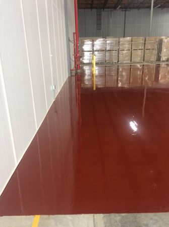 A warehouse scene with a high-gloss red floor reflecting the environment. To the left is a white wall with electrical conduit and a fire extinguisher, leading to a red column. In the background, stacked pallets of goods are visible. A yellow line on the floor suggests safety or organizational guidelines within the facility.