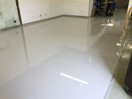 A newly coated glossy white floor transitions to an older, gray concrete surface near the walls, reflecting the bright overhead lighting. The room appears to be under renovation, indicated by unfinished walls and electrical outlets. A utility cart and a shop vacuum stand to the right, suggesting ongoing work in a commercial or industrial setting.