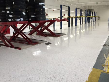 A view of a well-maintained vehicle service workshop with a shiny, speckled floor reflecting the bright lighting. To the left, a stack of black tires is placed on a red scissor lift. The area features several blue car lifts in the background, and the floor is marked with yellow safety lines near a metal drain cover, suggesting a focus on order and safety in the workspace.