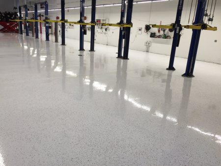 A glossy, speckled floor of a vehicle service area reflecting the lighting above. Multiple blue car lifts are evenly spaced throughout, with vehicles raised on some. The walls are white, and various automotive tools and equipment are mounted along the back wall, indicating a professional automotive service environment.