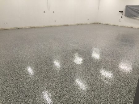 A spacious room with a speckled grey epoxy floor that reflects overhead lighting. White walls with electrical outlets and a baseboard run around the perimeter. A plastic sheet partially covers a segment of the wall on the right side, suggesting an area under construction or renovation.