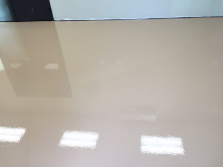 A glossy beige floor with reflections of fluorescent lights visible on its surface, showing a detail of the interior of a modern building. The edge of a black baseboard is visible at the top of the image, indicating a wall-floor junction.