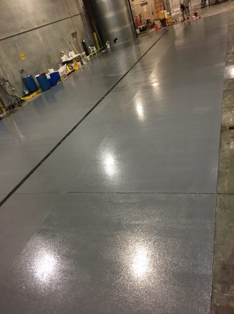 Diagonal perspective of a vast industrial floor with a smooth, light gray epoxy finish, showing a reflective sheen. The flooring extends towards a back area with various cleaning supplies and blue drums lined against the concrete wall, indicating a working or storage area.