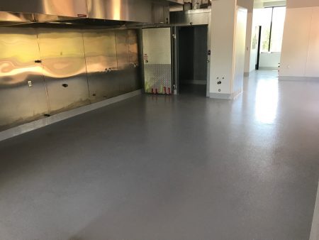 Spacious commercial kitchen area with shiny gray epoxy flooring reflecting overhead lights. The room includes stainless steel countertops against the wall, a metal door to a walk-in refrigerator, and an industrial fire extinguisher set. There's an entrance to another room on the right, showing a glimpse of a green space outside.