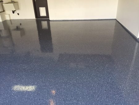 A spacious room with a highly reflective navy blue epoxy floor speckled with tiny white and light blue flecks. The glossy surface mirrors the white walls, a brown door, and a utility sink on the left side. The bright reflections and the spotless condition of the floor give the room a sleek and professional appearance, common in automotive garages or high-end workshops.