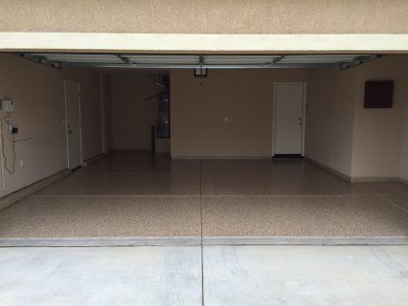 View from the driveway into a double garage with tan speckled epoxy flooring. The garage is open, revealing a clean, uncluttered space with two white doors on the back wall, a water heater to the right, and an electrical box on the left wall. The ceiling texture and walls are painted in a matching tan color, providing a uniform and tidy appearance.