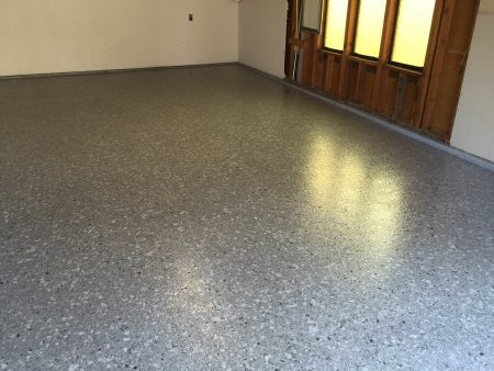 A room with a gray speckled epoxy floor casting a light sheen. Sunlight streams in through textured glass windows set in a wooden frame on the right wall, creating bright patterns on the floor. The left wall is white with a visible electrical outlet and a bare corner. This image suggests a residential space with a blend of warmth and functionality.