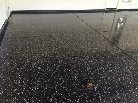 ALT text: "A reflective black epoxy floor with a dense pattern of small white speckles. The high gloss finish of the floor creates mirror-like reflections of the white walls and a door within the room. The baseboard is also black, blending seamlessly with the flooring. This sleek and polished surface suggests a modern and clean space, possibly in a residential or commercial setting.