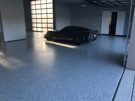 Late afternoon sunlight filters into a garage with a textured grey epoxy floor, highlighting a dark grey sports car with distinctive red brake calipers. The garage is spacious and uncluttered, with the car positioned in the center, facing an open garage door. The light creates a serene atmosphere, emphasizing the sleek design of the car and the clean, practical space.