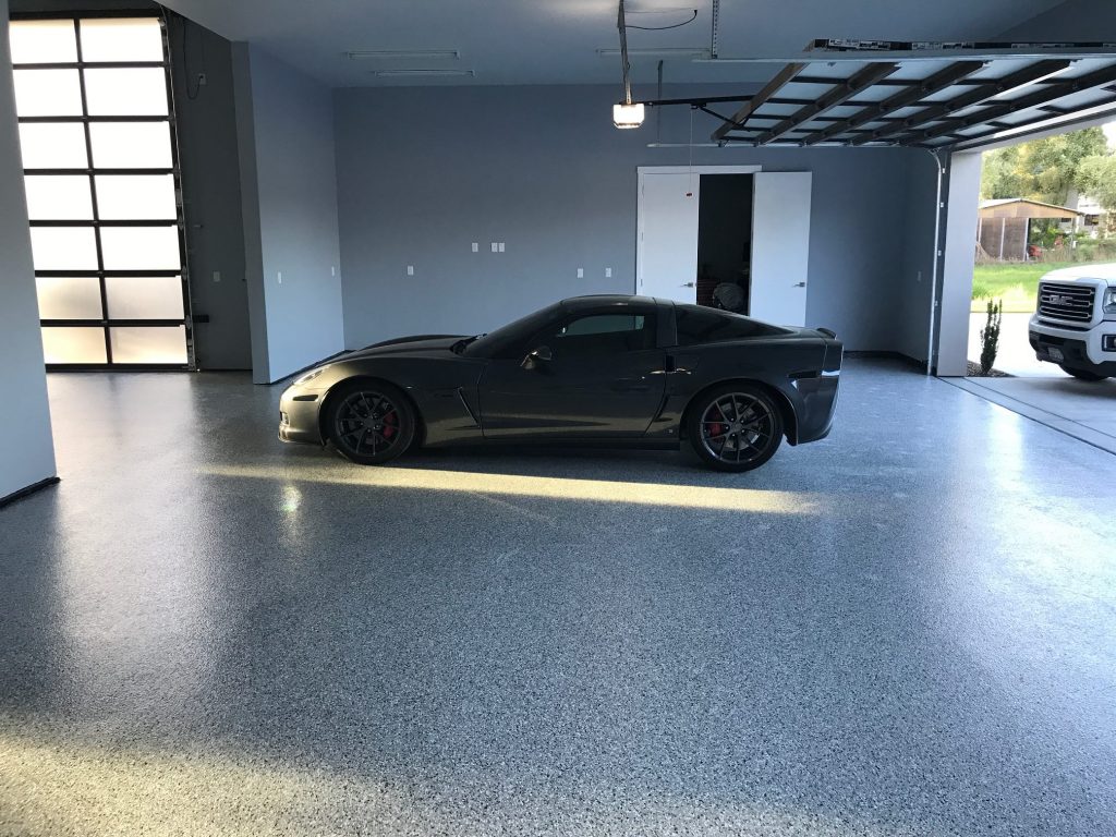 A spacious garage with a speckled grey epoxy floor with top coat, housing a sleek dark sports car with red brake calipers. Natural light streams in through the frosted glass of the garage door, casting a warm glow on the vehicle and floor. The clean and organized space conveys a modern look with a touch of luxury, emphasized by the high-end car parked inside.