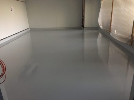 A room with a light grey epoxy floor that has a subtle reflective sheen, complementing the pale walls and creating a clean, minimalistic look. The room is partially lit, with shadows near the edges and a protective plastic sheeting visible, hinting at ongoing work or recent renovations. The image conveys a sense of calm and spaciousness, often associated with modern industrial or commercial interiors.