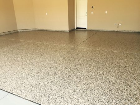 An empty room with speckled terrazzo flooring and beige walls. There is a white door on the far wall with a light switch to its right and two electrical outlets on adjacent walls. The room has a baseboard that matches the floor, providing a seamless transition between the wall and floor.
