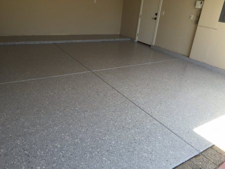 A spacious garage interior with a clean, freshly applied epoxy floor coating in a speckled grey finish. The floor is divided by expansion joints creating large rectangular sections. The walls are painted beige, and two closed white doors are visible, one with an electronic keypad entry. Sunlight streams in from the open garage entrance, highlighting the floor's glossy texture.