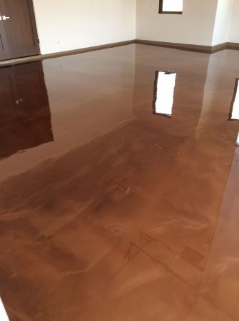 A spacious room with a highly reflective caramel-colored epoxy floor that mirrors the windows and the ceiling's details. The room has neutral-colored walls with dark baseboards and a door on the left. The high gloss finish of the floor gives the space a sleek and modern look, suggesting recent construction or renovation in a residential or commercial setting.