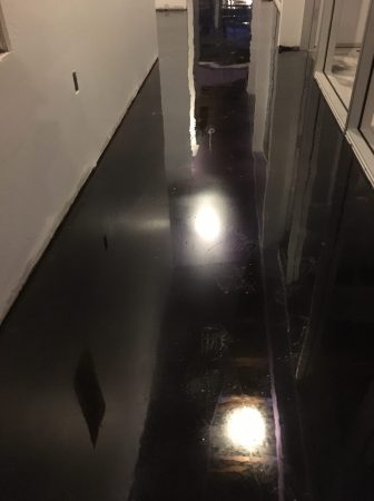 A narrow corridor with shiny black flooring that reflects the overhead lighting. The corridor is flanked by a white wall on the left and a series of glass panes on the right, revealing another dark room or outside area. The reflection on the floor creates a striking contrast with the dark surface, suggesting the floor may be wet or polished to a high shine. Footprints and other marks are slightly visible on the floor's surface.