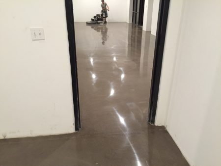 View from a doorway showing a person operating a floor cleaning machine in a brightly lit corridor with reflective flooring. The walls are white, and the corridor leads to a series of doorways. An electrical outlet is visible on the wall to the left, indicating the setting is likely a commercial or institutional building. The reflection of the lights on the floor highlights the cleanliness and maintenance of the space.