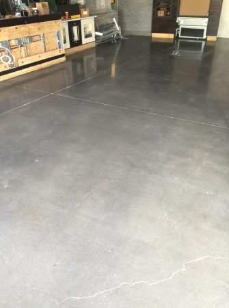 Interior view showcasing a matte gray concrete floor with subtle crack patterns. A wooden service counter with construction materials on it is in the background, indicating an unfinished or under-renovation space. To the right, metal shelving and a portable heater are visible, with a dark tiled wall partially seen in the background. The space suggests a commercial setting, likely a shop or a café in the process of being set up or refurbished.