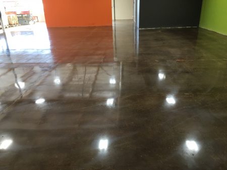 A view of a spacious room with a polished concrete floor, reflecting the ceiling lights. The walls are painted in bold orange on the left and a combination of black and lime green on the right, suggesting a vibrant and modern setting. The space is likely part of a commercial or public building, characterized by its open floor plan and the visibility of an adjacent area through a doorway on the left, where merchandise and shelving units can be glimpsed.