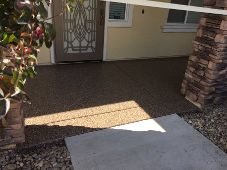 Residential entrance with a textured, speckled concrete walkway leading up to a door with a decorative security screen. The entry is flanked by stone column accents and a small window to the right, with a landscaped area of pebbles and shrubs to the left.