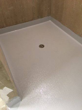 Corner shower base with a freshly applied non-slip coating, featuring a central round drain. The walls around the base are unfinished with visible plywood backing.