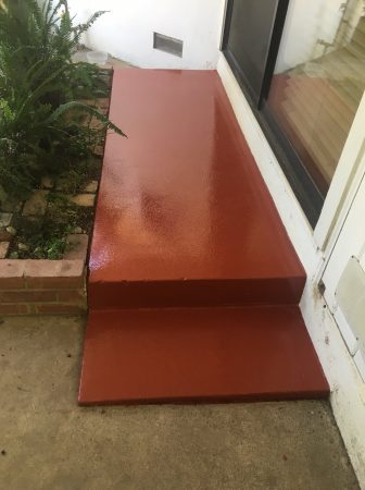 A freshly painted, terracotta-red concrete doorstep leading to a white door, contrasting with the surrounding concrete walkway. To the left is a garden bed with a variety of green ferns and a brick border, adding a touch of nature to the entrance of the building.