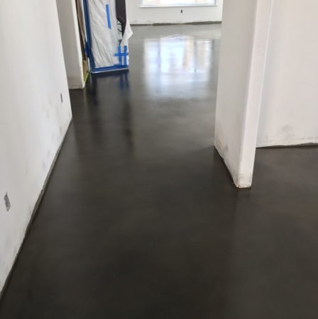 Narrow hallway with a dark, polished concrete floor leading to a brighter space with natural light, featuring walls with some scuff marks and a doorway covered with plastic sheeting, suggestive of an area under maintenance or renovation.