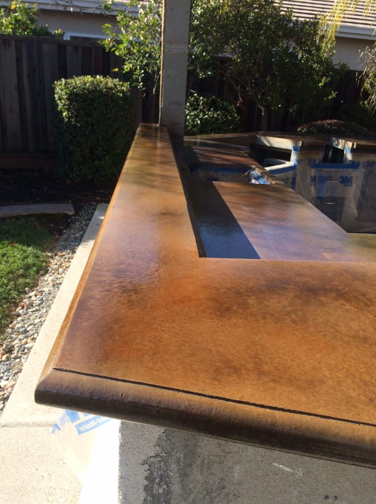An outdoor countertop with a rich, rust-colored concrete stain is featured in a garden setting, with construction materials and protective plastic sheeting in the background, indicating ongoing work on the property.