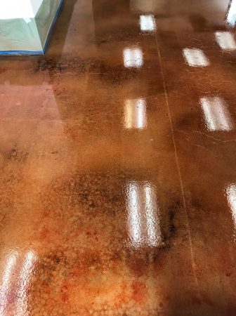 Close-up of a polished concrete floor with a rich terracotta stain and scattered light reflections, showcasing variations in hue and the high-gloss finish