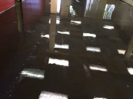 Reflective, wet-looking floor inside a building, with square light reflections and a few footprints scattered across the surface, between columns and under ambient light coming from windows.