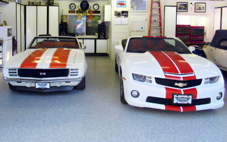 Two Chevrolet Camaros inside a brightly lit garage, facing forward. On the left is a classic 1969 Camaro SS with white paint and orange racing stripes, while on the right is a modern white Camaro with red stripes. The garage is well-organized with shelves, speakers, and automotive decor.