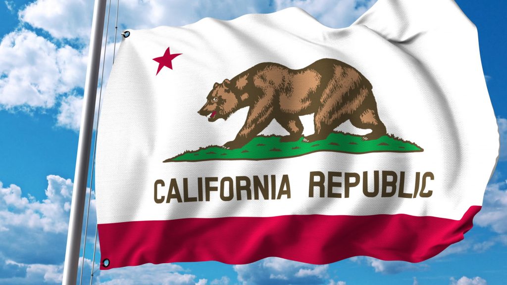 The state flag of California waving against a backdrop of a blue sky with scattered clouds. The flag features a central image of a grizzly bear walking on a patch of green grass, with a red star above the bear and the words 'California Republic' below. A red stripe runs along the bottom of the flag.