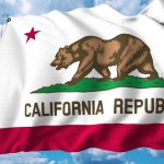 The state flag of California waving against a backdrop of a blue sky with scattered clouds. The flag features a central image of a grizzly bear walking on a patch of green grass, with a red star above the bear and the words 'California Republic' below. A red stripe runs along the bottom of the flag.