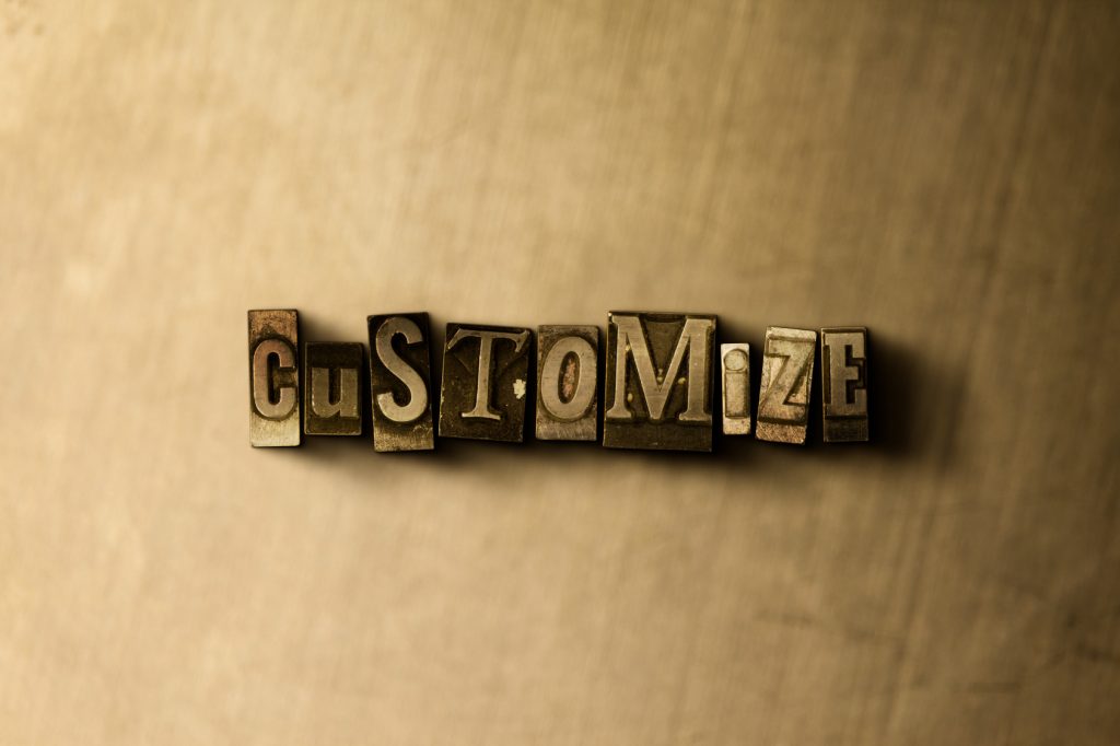 The word 'CUSTOMIZE' spelled out in vintage metal letterpress printing blocks, centered and casting a shadow on a textured beige background.