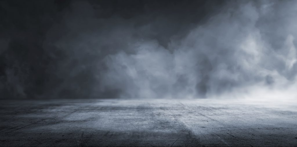 A dramatic, expansive view of a dark, textured concrete floor leading into a dense, misty atmosphere. The foreboding clouds above suggest a sense of uncertainty or suspense, possibly representing a scene from a thriller or horror genre. The image could evoke a metaphor for facing the unknown or entering into a challenging situation.