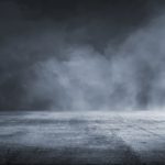 A dramatic, expansive view of a dark, textured concrete floor leading into a dense, misty atmosphere. The foreboding clouds above suggest a sense of uncertainty or suspense, possibly representing a scene from a thriller or horror genre. The image could evoke a metaphor for facing the unknown or entering into a challenging situation.