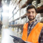 A confident male warehouse worker in a high-visibility orange vest stands in an aisle between tall shelves stocked with goods. He holds a clipboard, possibly taking inventory or managing orders. The large warehouse setting indicates an industrial or commercial environment, and the worker's pleasant expression suggests a positive workplace.