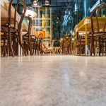 A ground-level view inside a modern cafe with an array of wooden chairs and tables. The furniture is neatly stacked, indicating the cafe is either closed or not yet open for customers. The polished stone floor reflects the ambient lighting, contributing to the establishment's clean and inviting atmosphere. The glass walls provide a glimpse of the urban setting outside.