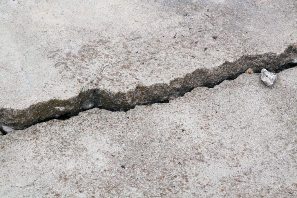 Close-up view of a jagged crack running through a concrete surface. The crack shows signs of aging and weathering, with small debris accumulated within. This image could indicate structural damage or wear, commonly seen in pavements, sidewalks, or other concrete structures.