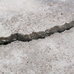Close-up view of a jagged crack running through a concrete surface. The crack shows signs of aging and weathering, with small debris accumulated within. This image could indicate structural damage or wear, commonly seen in pavements, sidewalks, or other concrete structures.