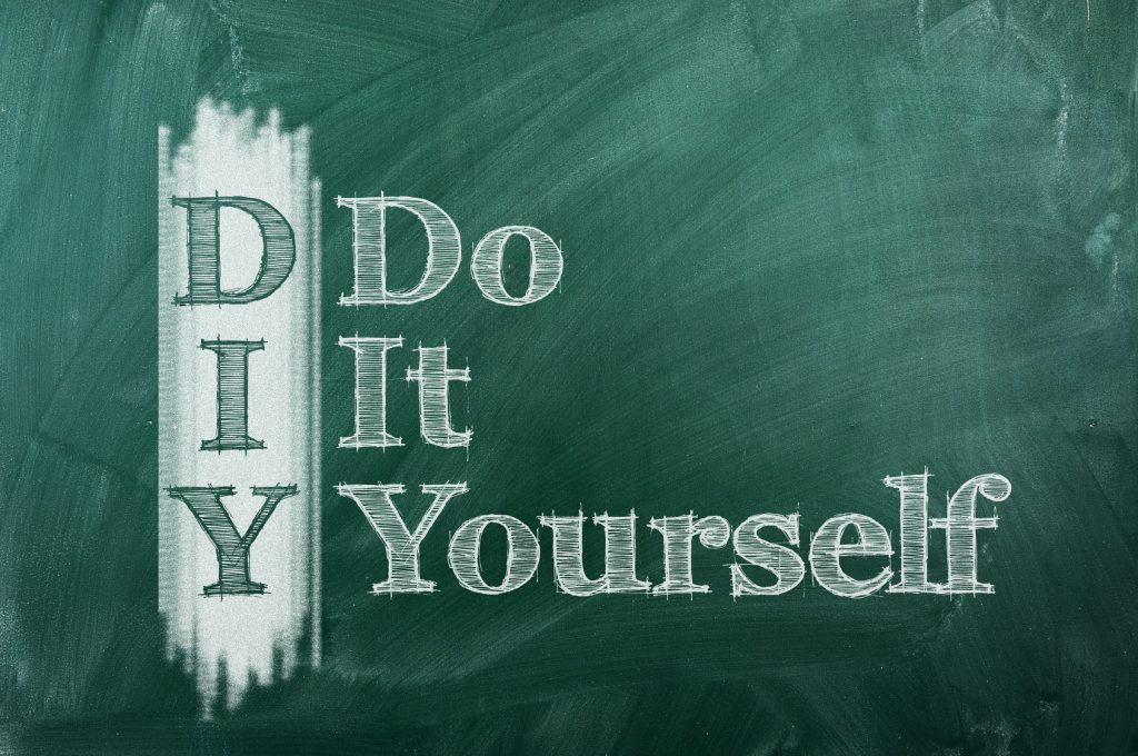 The acronym 'DIY' which stands for 'Do It Yourself,' is written in large white chalk letters on a dark green chalkboard. The first letters of each word are vertically aligned to form the acronym 'DIY' with the rest of the words written to the right, creating an instructional or motivational message often associated with crafting or home improvement projects.