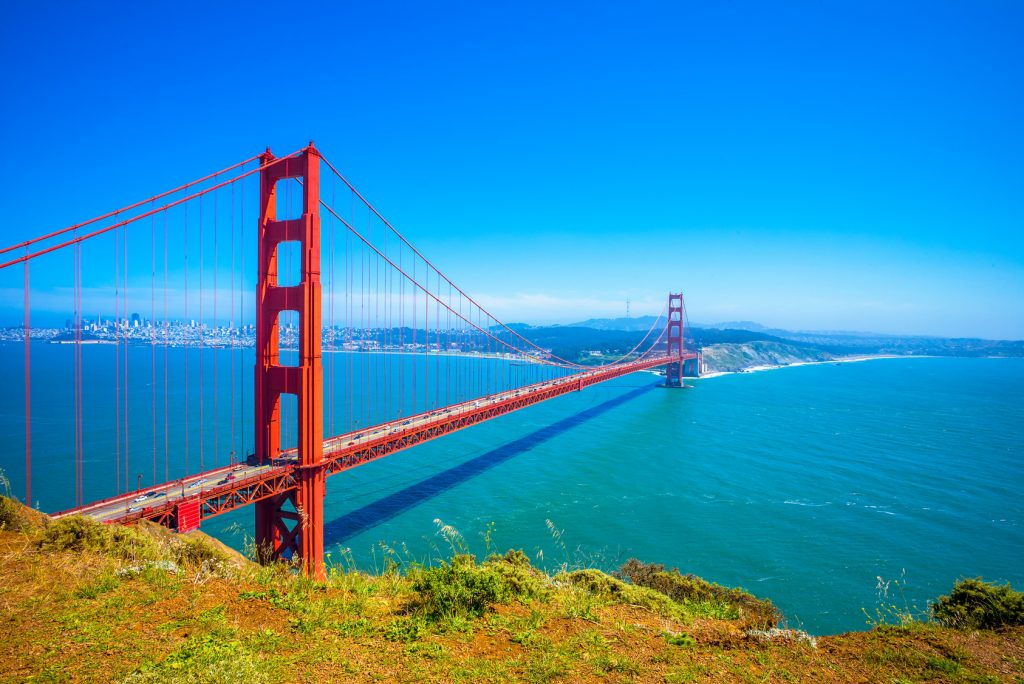 The iconic Golden Gate Bridge on a clear sunny day, with its striking International Orange color standing out against the blue sky and waters of San Francisco Bay. The bridge spans from the hilly, green landscape in the foreground to the urban skyline in the distance. This picturesque view captures the grandeur of this famous suspension bridge and its surroundings.