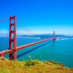 The iconic Golden Gate Bridge on a clear sunny day, with its striking International Orange color standing out against the blue sky and waters of San Francisco Bay. The bridge spans from the hilly, green landscape in the foreground to the urban skyline in the distance. This picturesque view captures the grandeur of this famous suspension bridge and its surroundings.